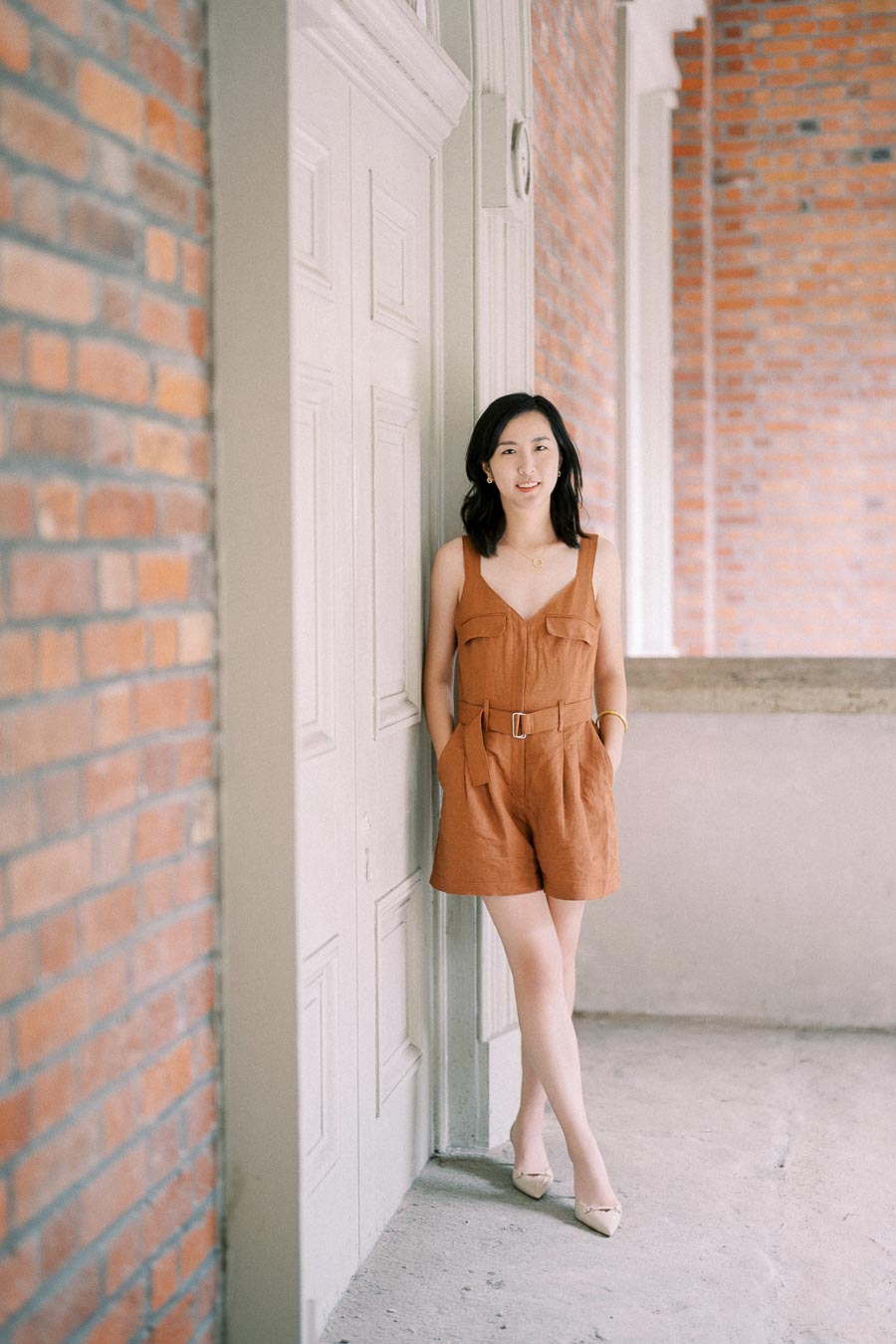 A woman in a brown romper stands against a white door, with a brick wall in the background, creating a stylish and relaxed atmosphere.