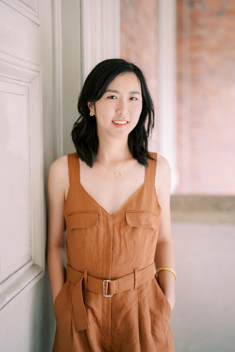 A woman wearing a brown sleeveless jumpsuit stands against a white wall, smiling softly.