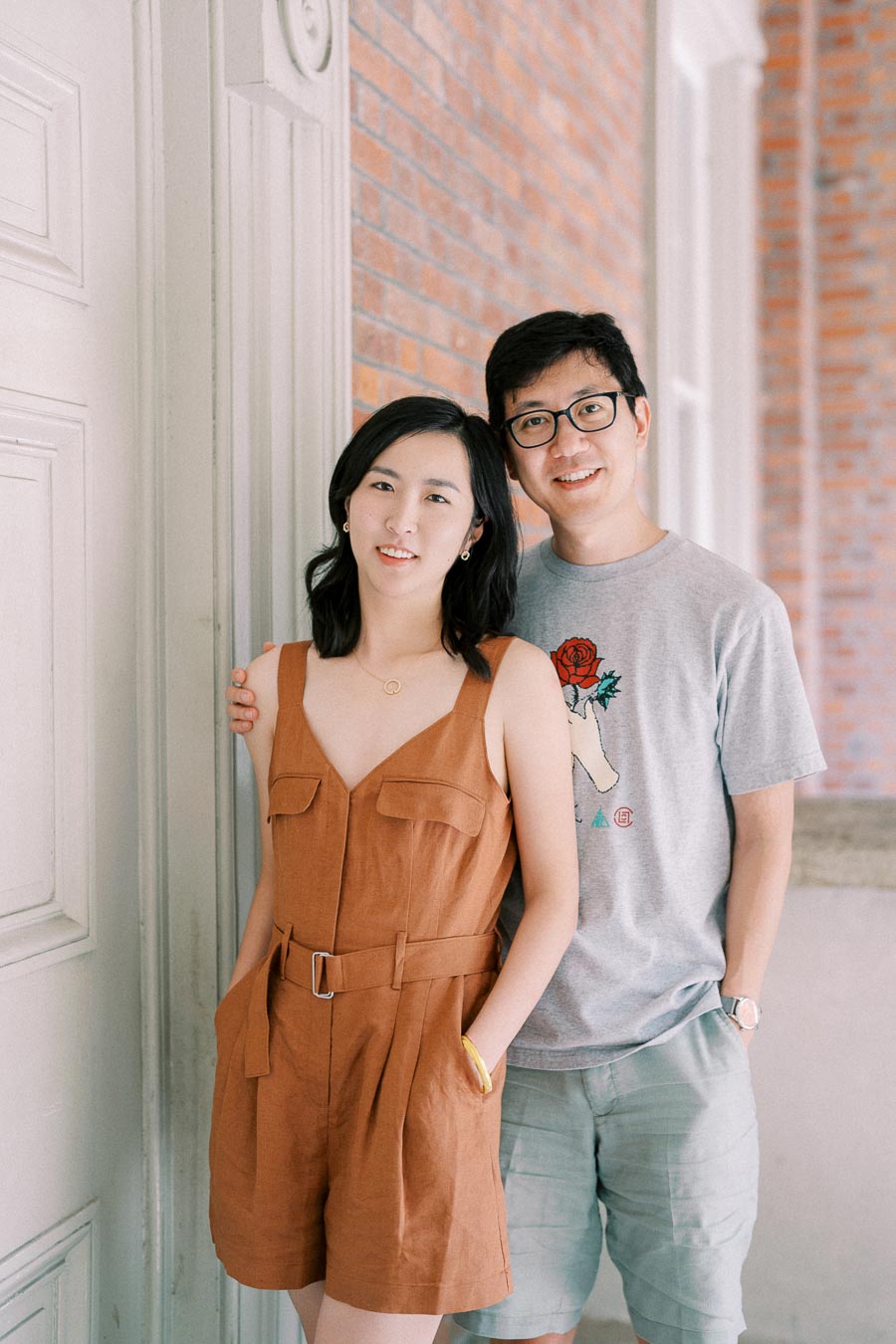 A couple poses together indoors, with the woman wearing a brown dress and the man in a gray t-shirt, standing against a brick wall and white door.