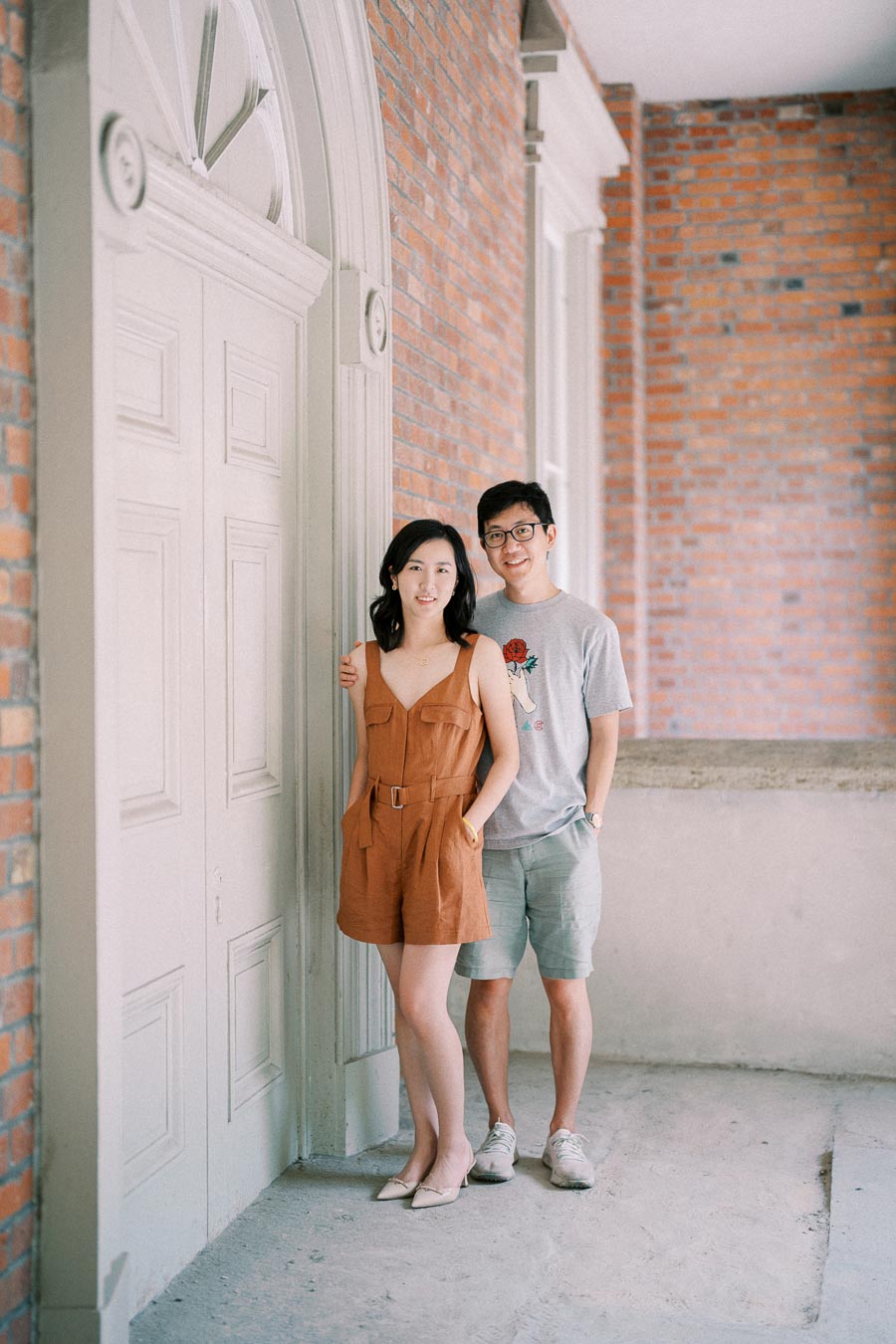 A couple poses in front of a white door with a brick wall background, showcasing a casual and relaxed atmosphere.