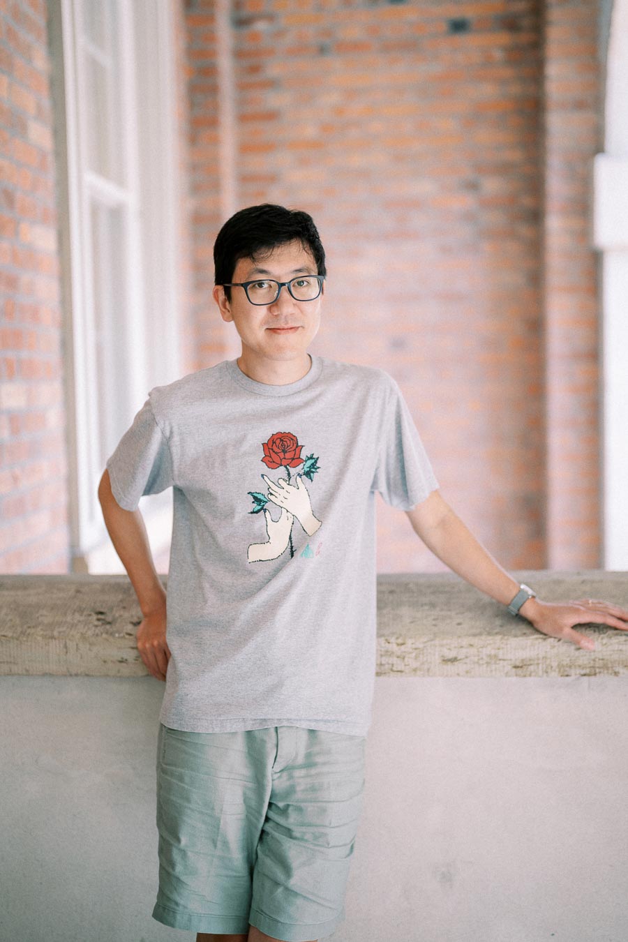 A person stands casually against a brick wall, wearing a gray t-shirt with a rose design and glasses.