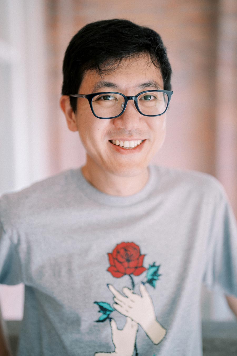 A person smiling while wearing glasses and a T-shirt featuring a graphic of hands holding a red rose.