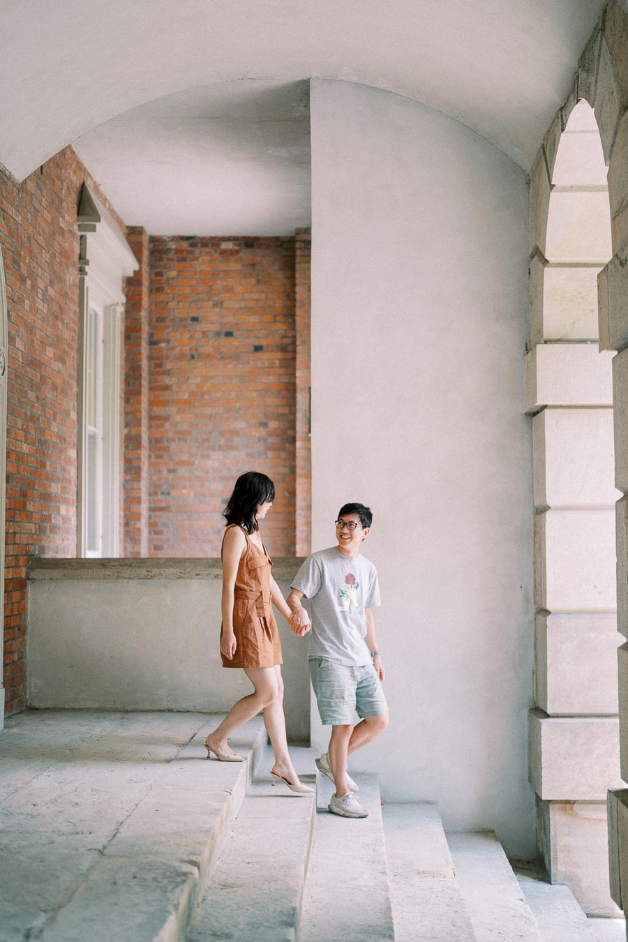 A couple walks hand in hand down the steps of a historic building with brick and stone architecture.