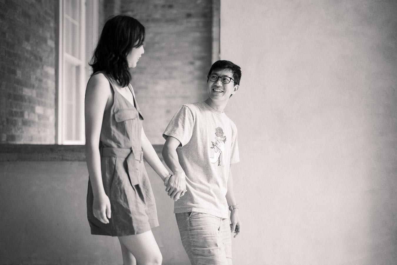 A couple shares a joyful moment, holding hands and smiling at each other in a black and white photograph.