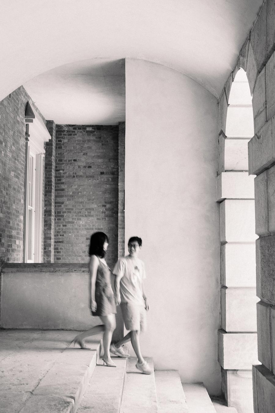 A black and white image captures two people walking down steps in an arched hallway, featuring brick and stone architecture.