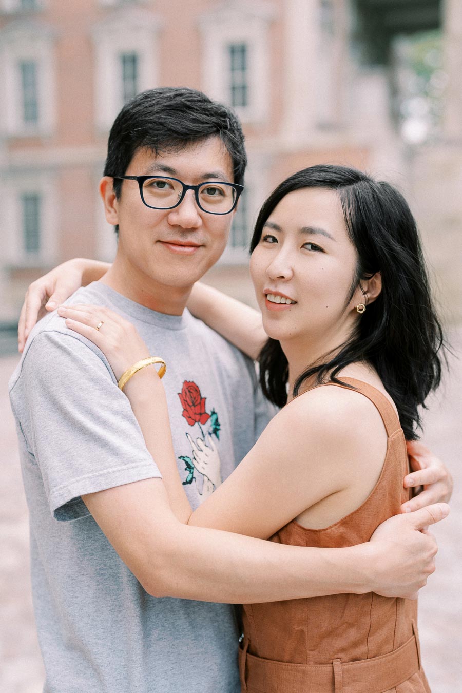 A couple embraces in front of a historic building, with the man wearing glasses and a graphic t-shirt, and the woman in a brown sleeveless dress.