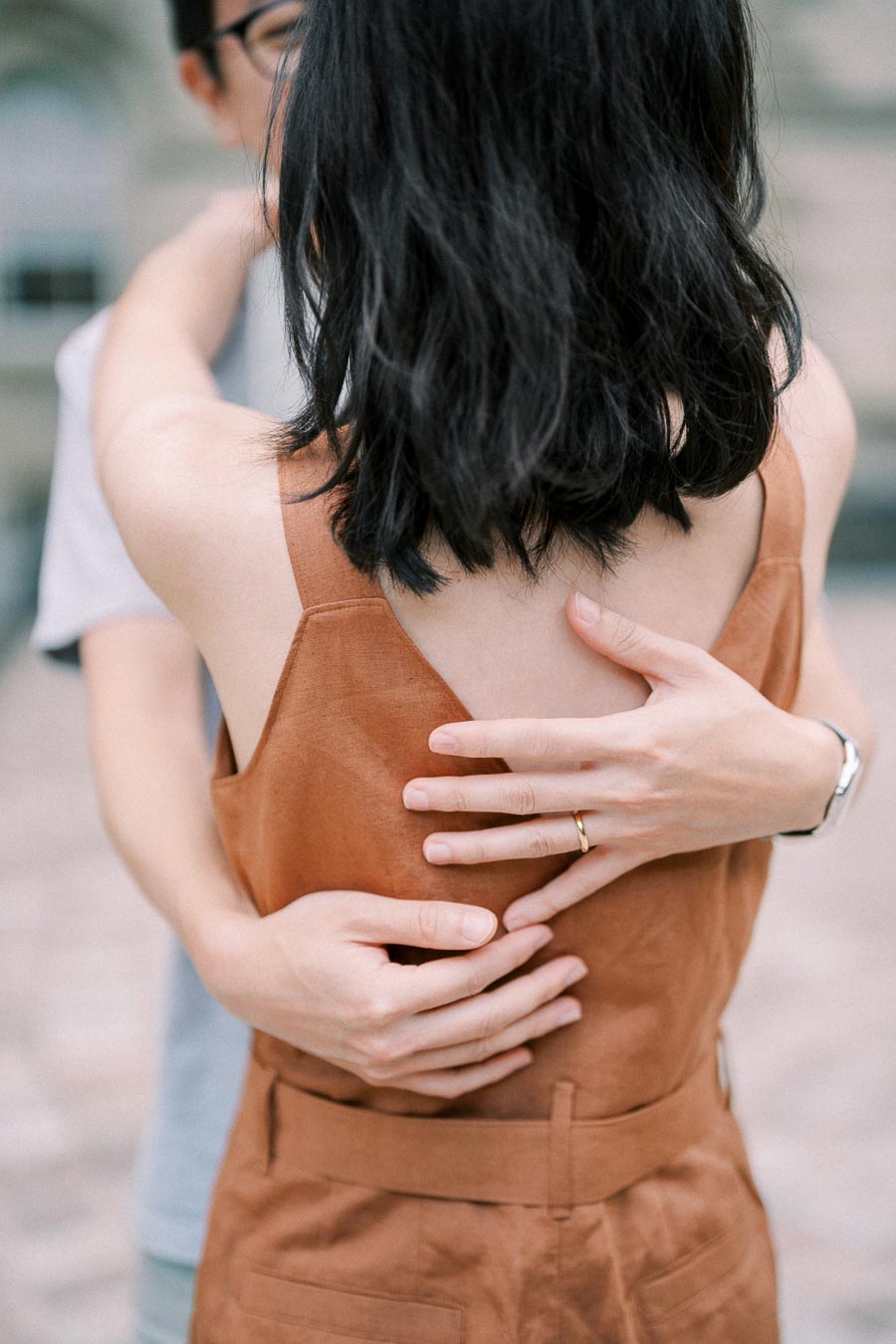 A close-up of a couple embracing, highlighting the woman's brown dress and the man's watch and ring.