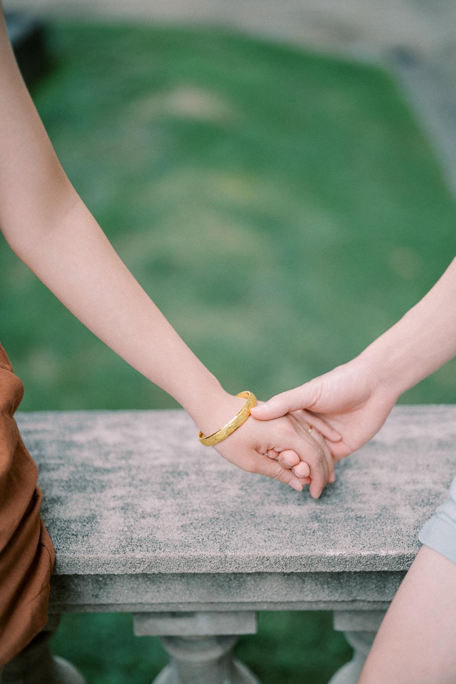 A close-up of two people holding hands over a stone bench, with one wearing a gold bracelet.