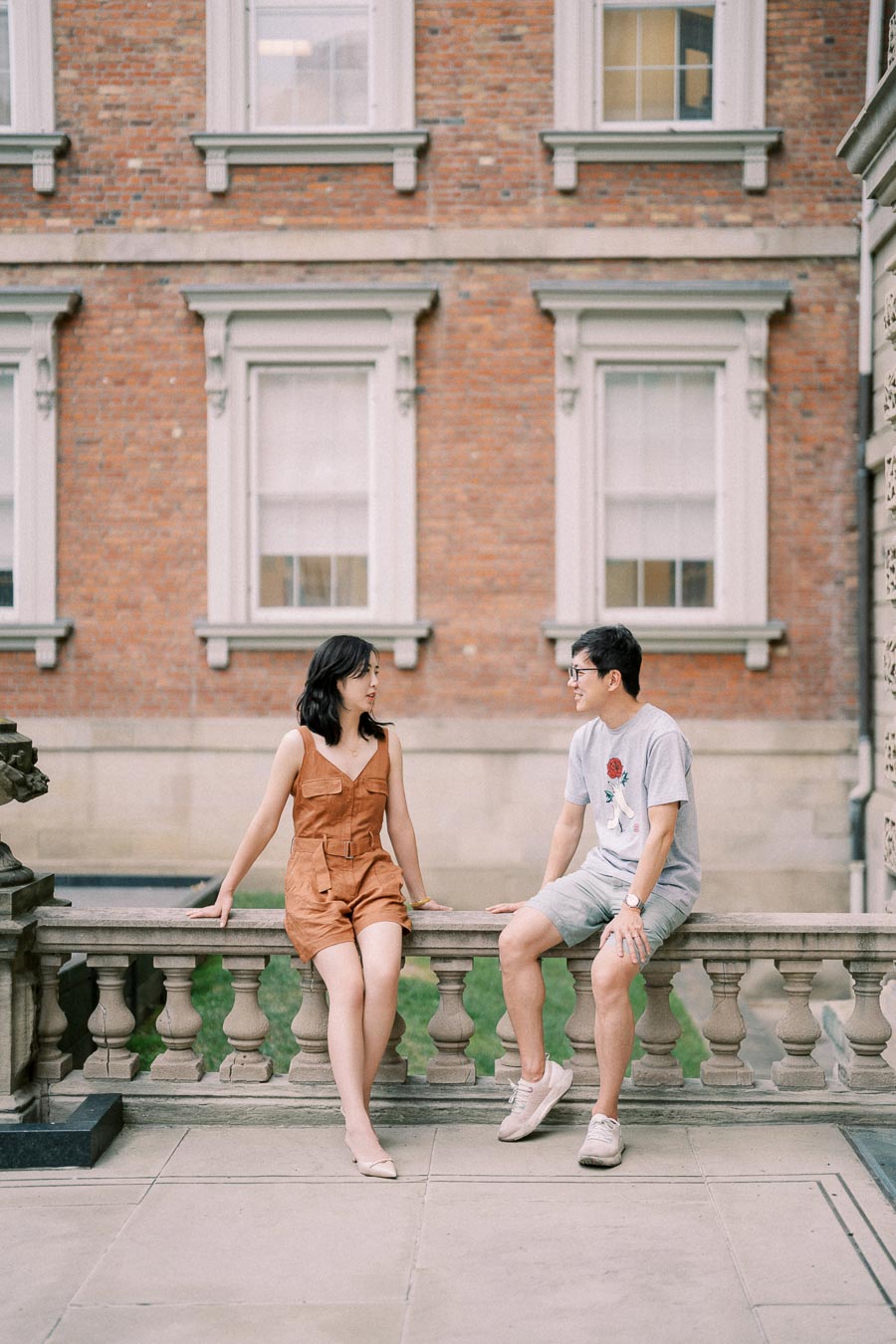 A man and woman sit on a stone railing in front of a historic brick building, engaged in conversation.