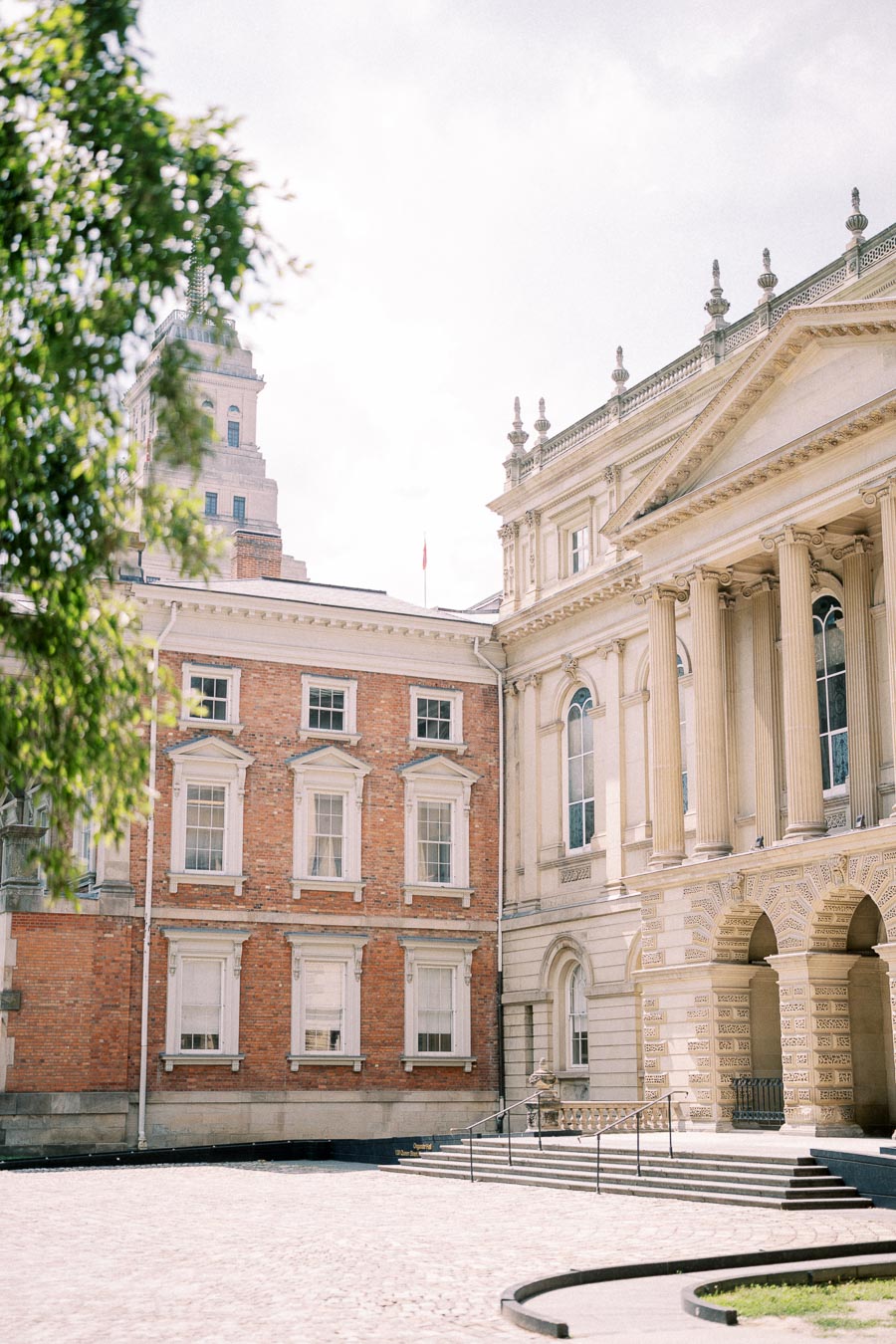A historic building featuring classical architecture with ornate columns and detailed stonework stands next to a red brick building. The scene is set under a bright, partly cloudy sky, with a tree partially visible in the foreground.
