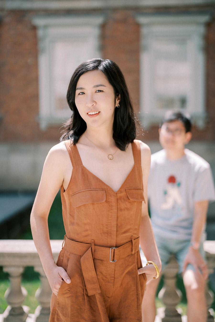 A woman in a brown sleeveless dress poses outdoors, with a blurred figure in the background.