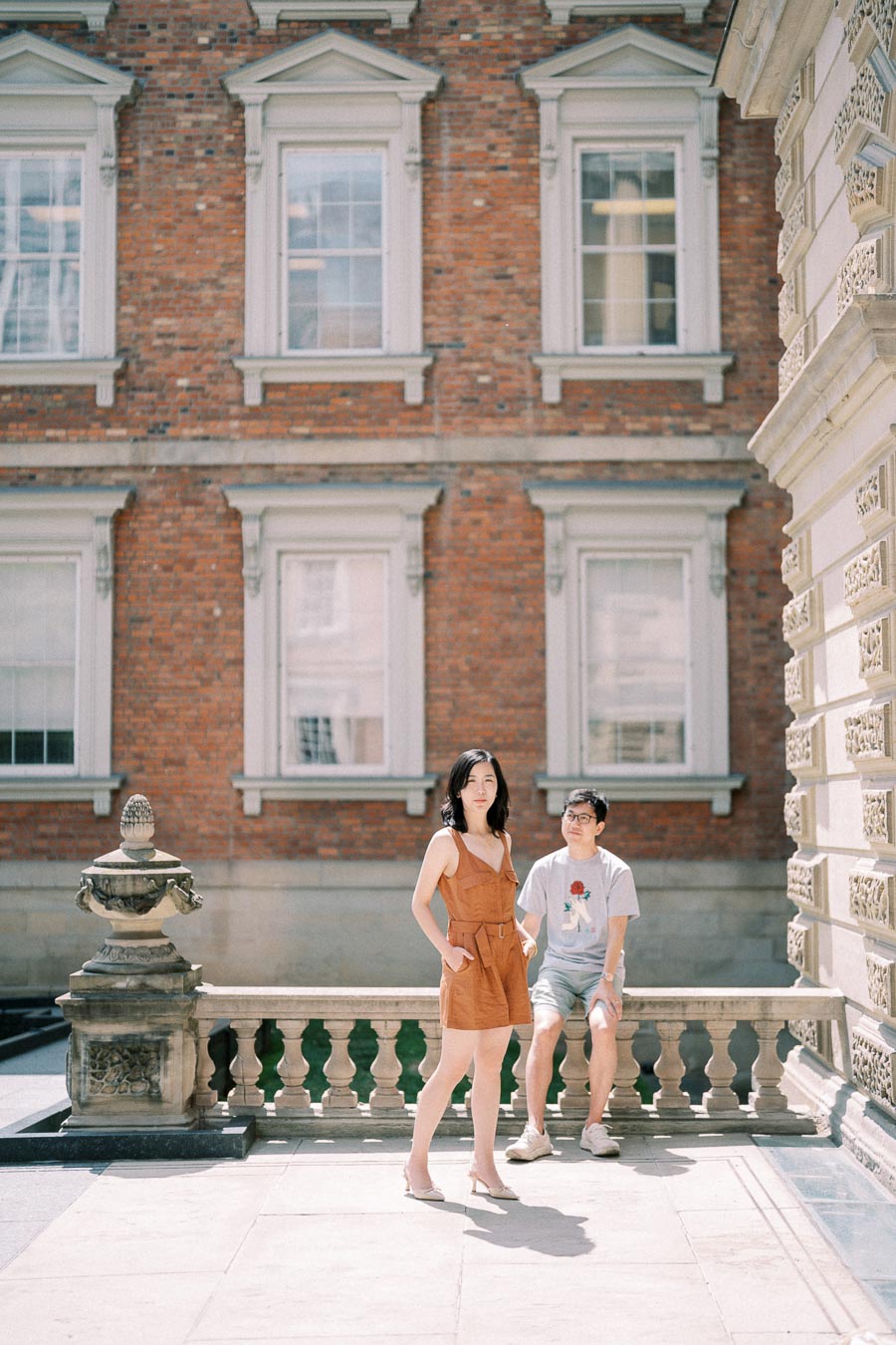 A woman poses in front of a historic brick building, while a man sits on a stone railing nearby.