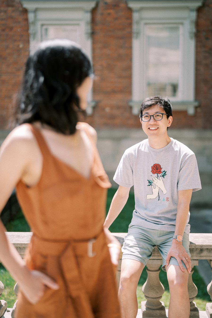 A casual outdoor interaction between two people, with one sitting on a stone railing and the other standing, set against a brick building backdrop.
