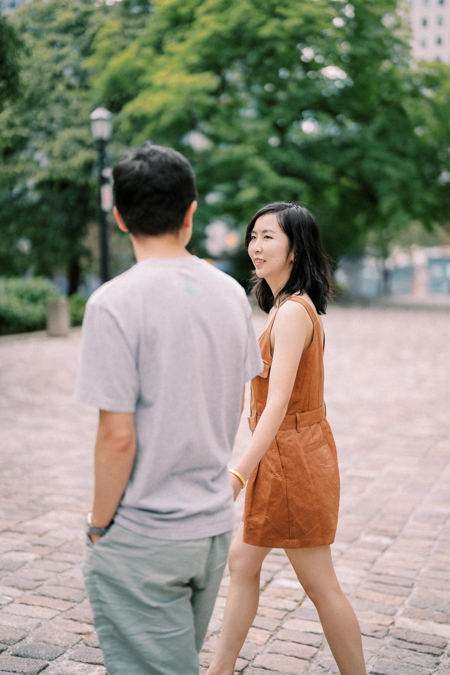A woman in a brown dress converses with a man in a gray t-shirt as they walk on a cobblestone path, surrounded by lush greenery.