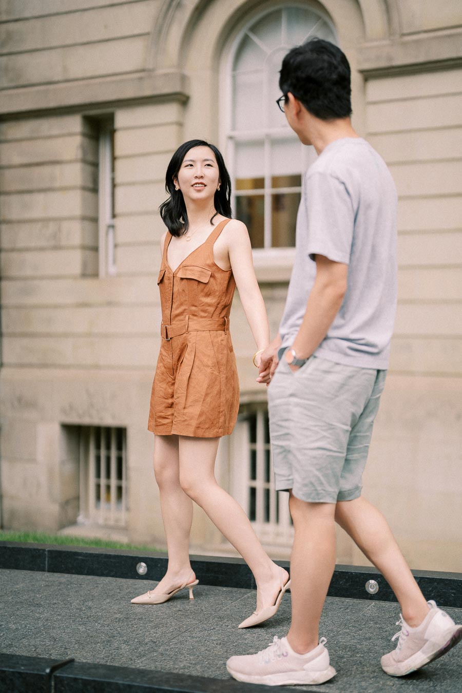 A couple walks hand in hand in front of a historic building, with the woman wearing a brown dress and the man in casual attire.