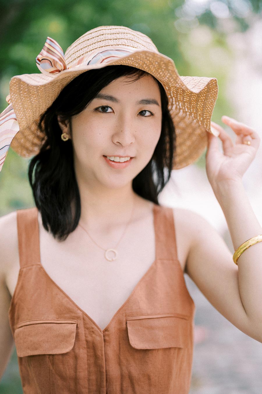 A woman smiles while wearing a straw hat with a ribbon and a brown sleeveless dress, accessorized with gold jewelry, in an outdoor setting.