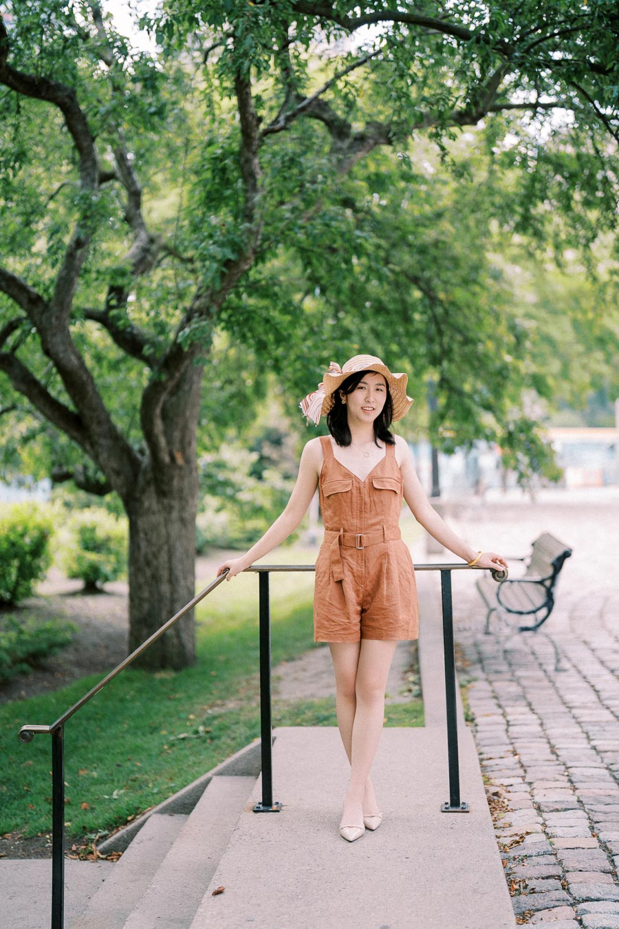 A woman wearing a brown romper and a wide-brimmed sun hat stands on a park pathway, surrounded by lush green trees and a bench in the background.