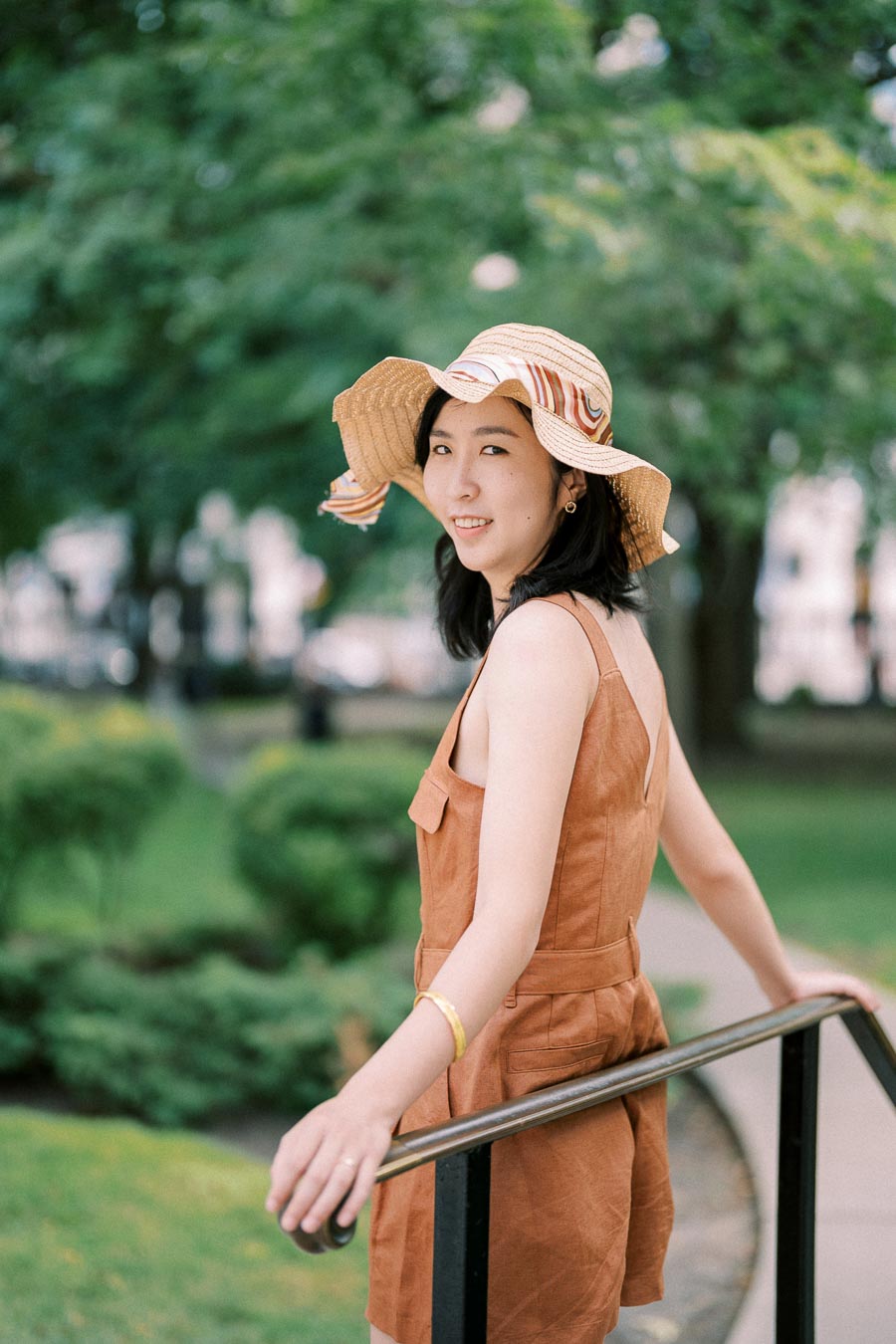 A woman wearing a brown dress and a straw hat stands by a railing in a lush green park, smiling at the camera.