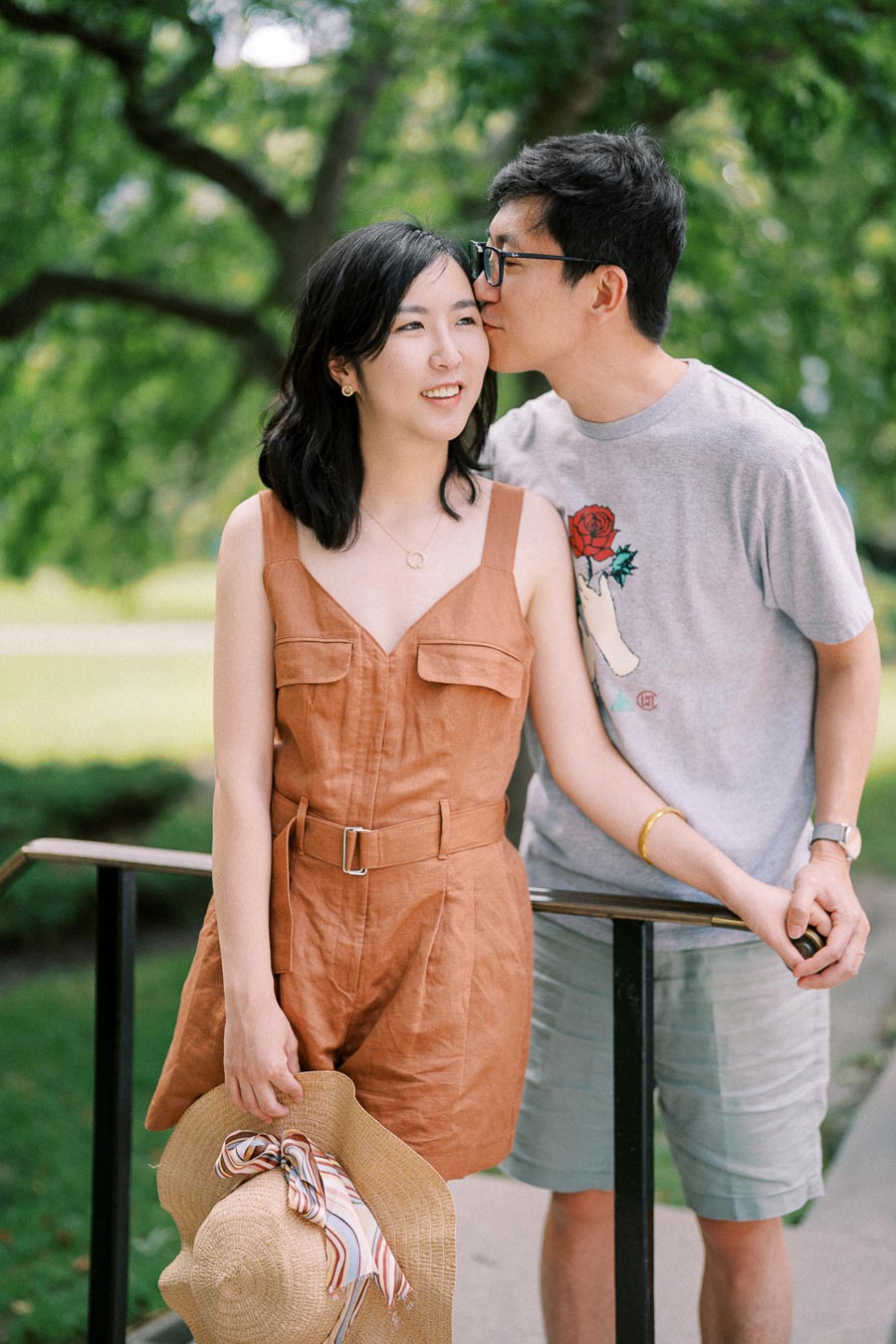 A couple shares a tender moment outdoors, with the man giving a gentle kiss on the woman's cheek as she holds a straw hat.