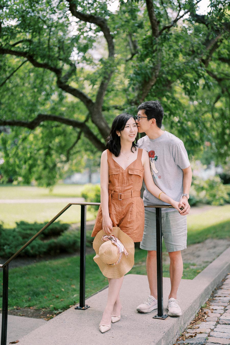 A couple enjoys a moment together in a lush green park, with the man whispering to the woman as she holds a straw hat.