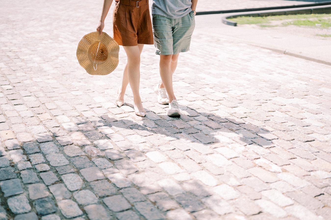 A couple strolls down a sunlit cobblestone street, with one person holding a straw hat.