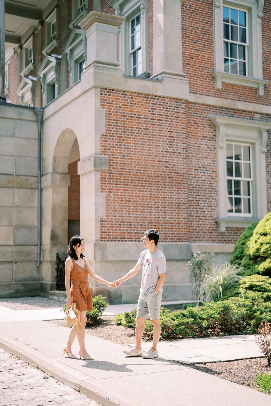 A couple holds hands in front of a historic brick building, enjoying a sunny day.