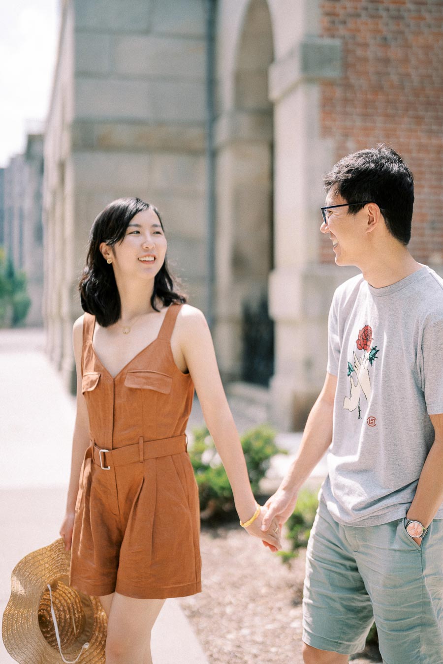 A couple enjoys a sunny day, holding hands and smiling as they walk near a brick building.