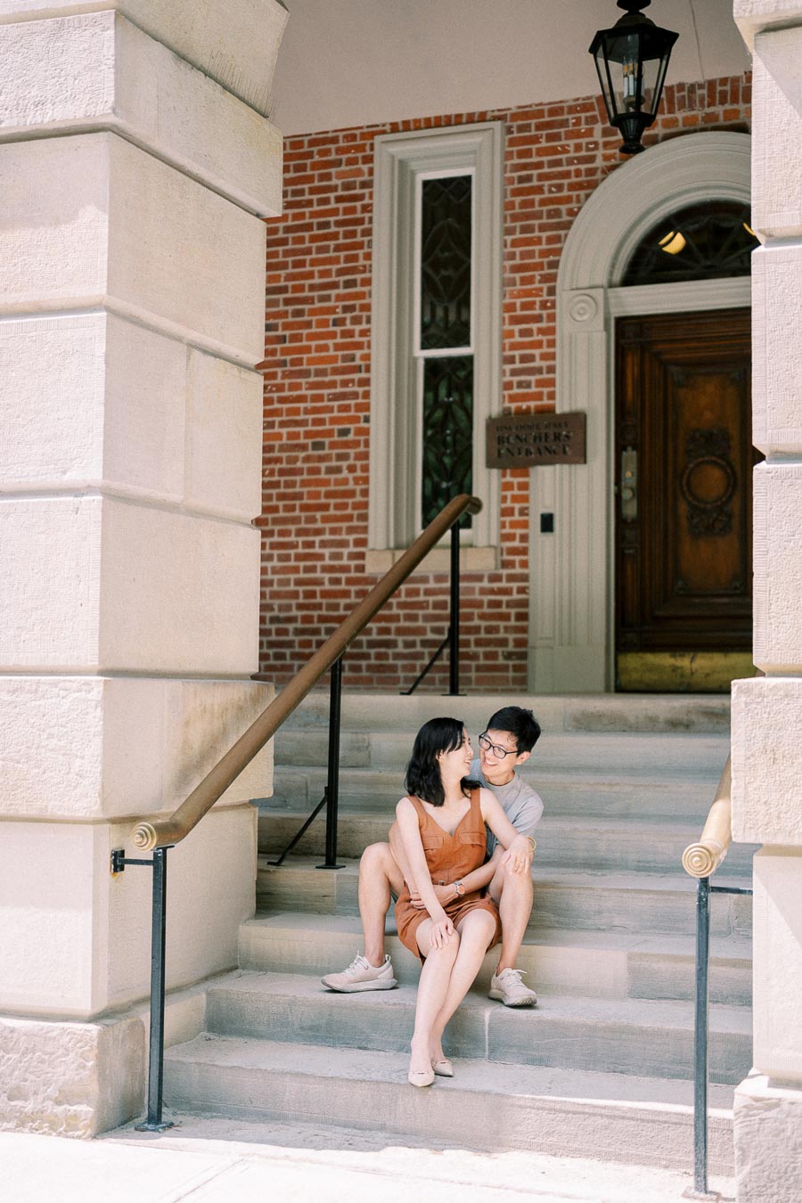 A couple sits affectionately on the steps of a historic brick building, enjoying a sunny day.