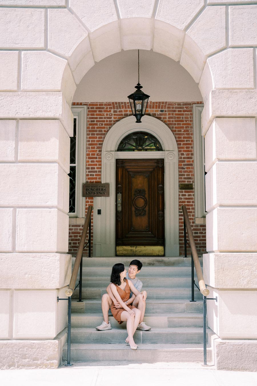 A couple sits on the steps of a brick building with an arched entrance, enjoying a sunny day.