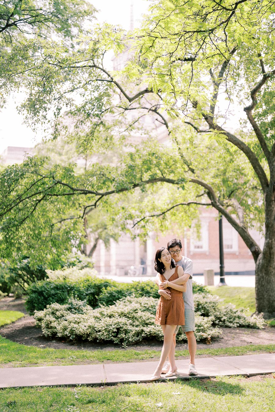 A couple enjoys a sunny day in a park, surrounded by lush greenery and a building in the background.