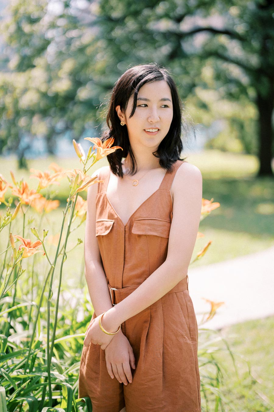 A woman stands in a park next to blooming orange lilies, wearing a brown dress and a gold bracelet.