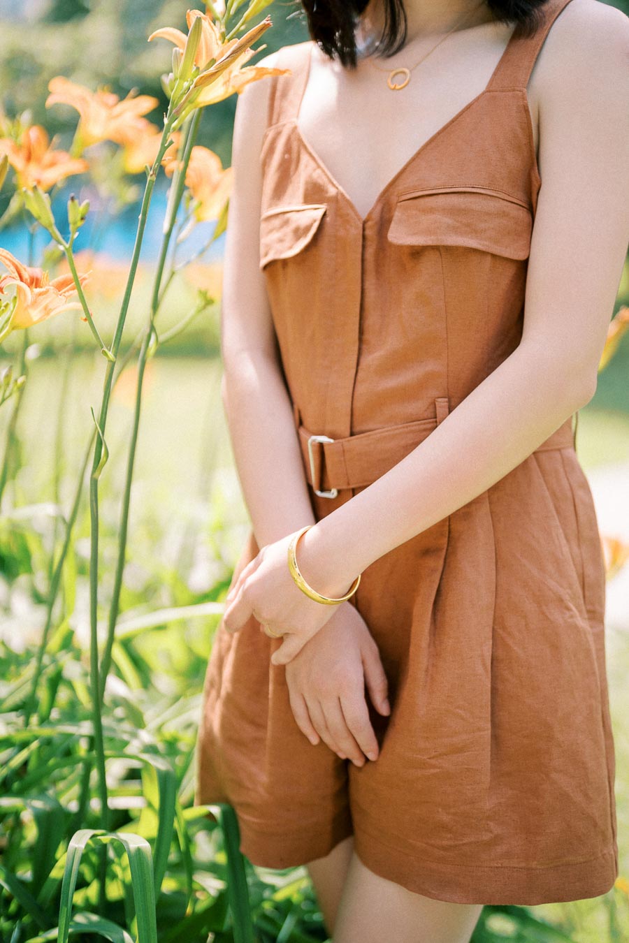 A woman in a brown dress stands in a garden, surrounded by blooming orange lilies. She is wearing a gold bracelet and necklace, with her hands gently clasped in front of her.