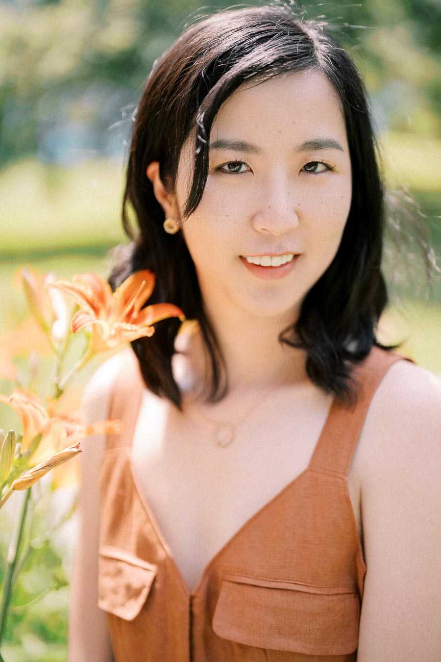 A woman smiles next to vibrant orange lilies in a sunny garden setting.