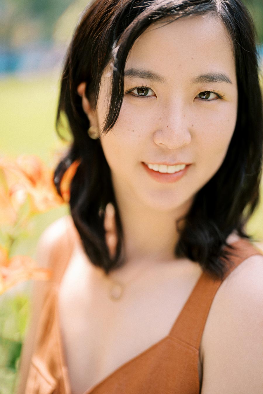 A woman with dark hair smiles warmly in a sunny outdoor setting, with orange flowers in the background.