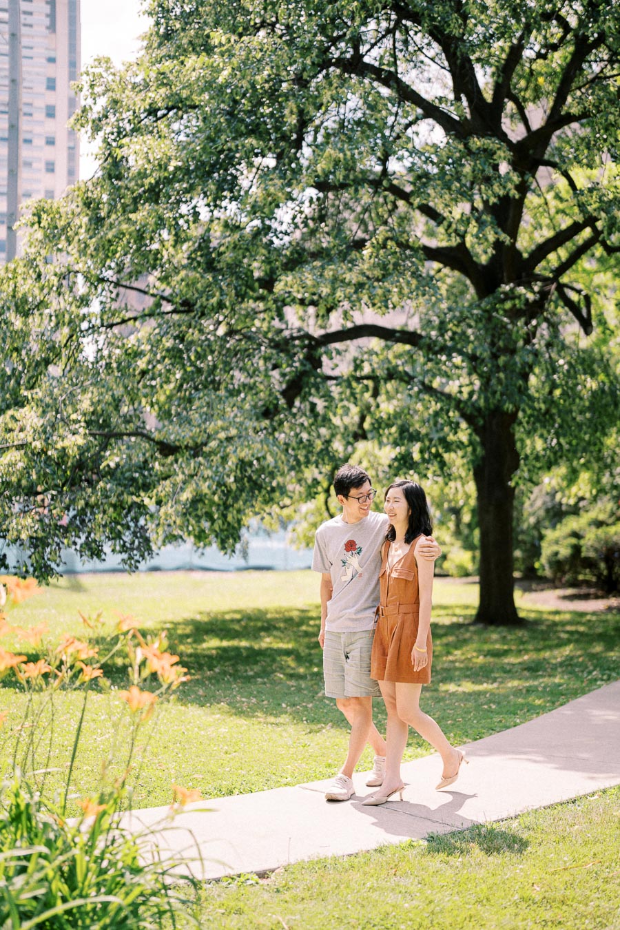 A couple enjoys a sunny walk in a lush park, surrounded by vibrant greenery and flowers.