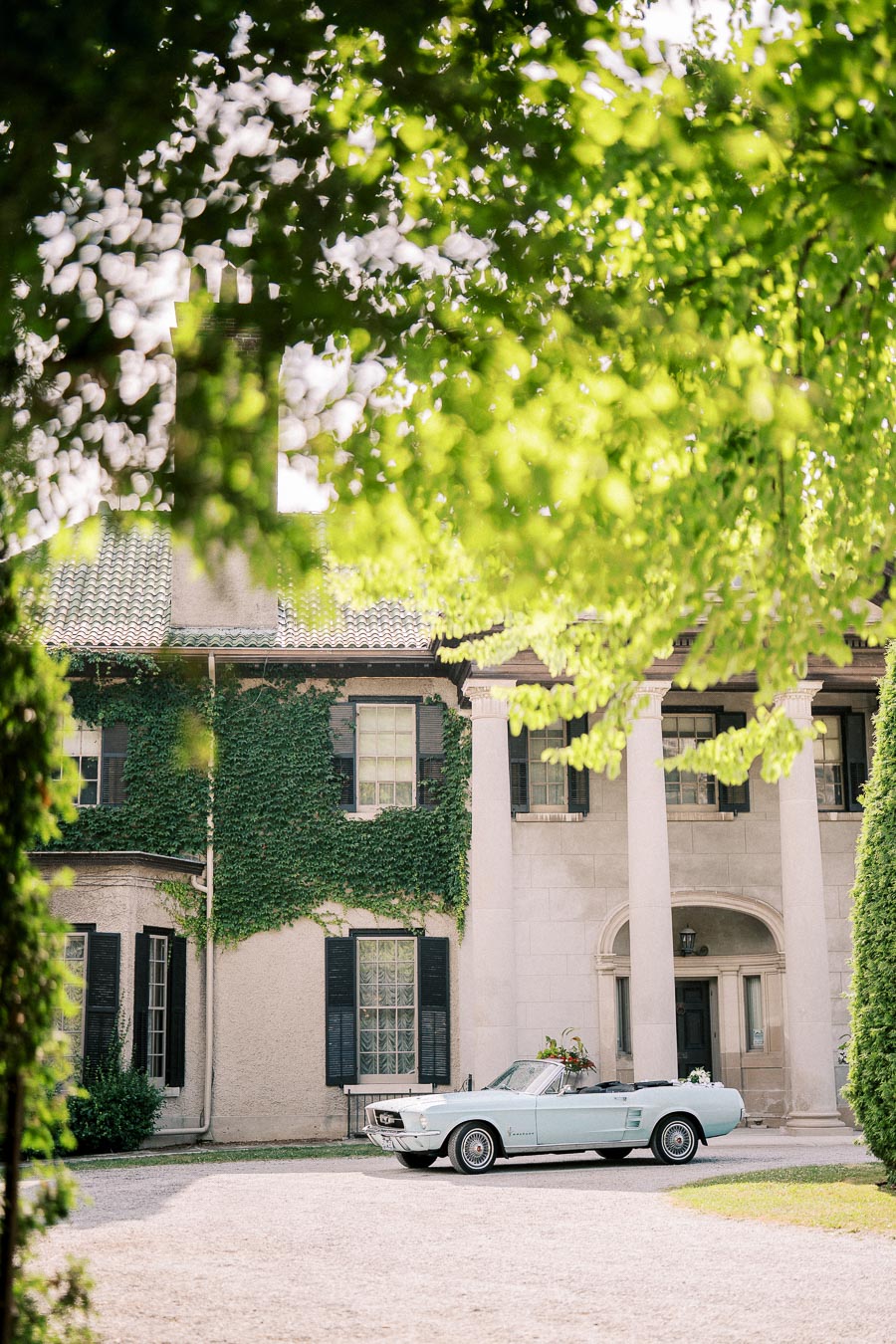 Vintage convertible parked in front of an elegant ivy-covered mansion with large pillars, surrounded by lush green foliage.