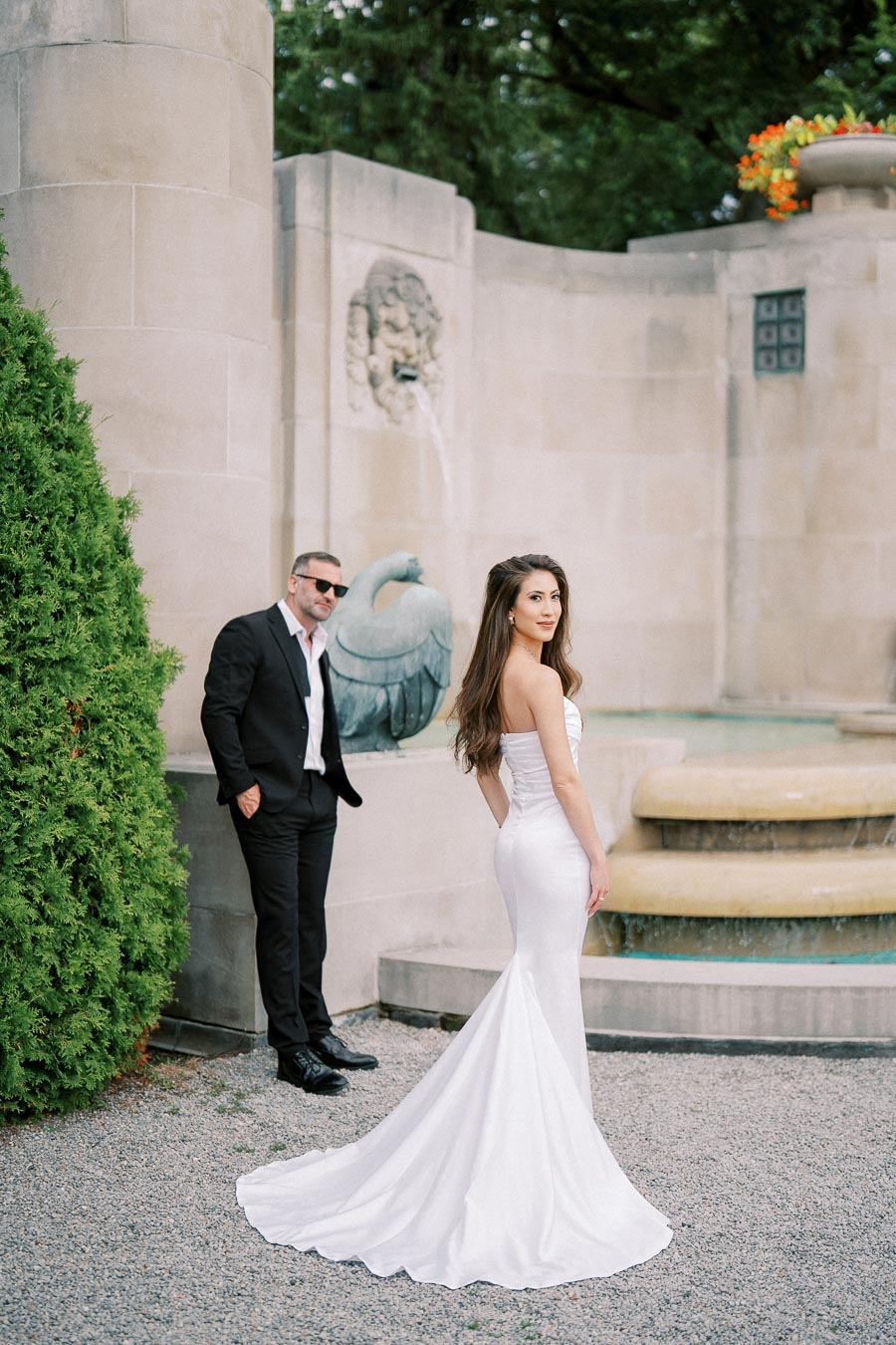 A bride in a strapless white gown poses near a stone fountain, with a groom in a black suit and sunglasses leaning against a wall adorned with greenery.
