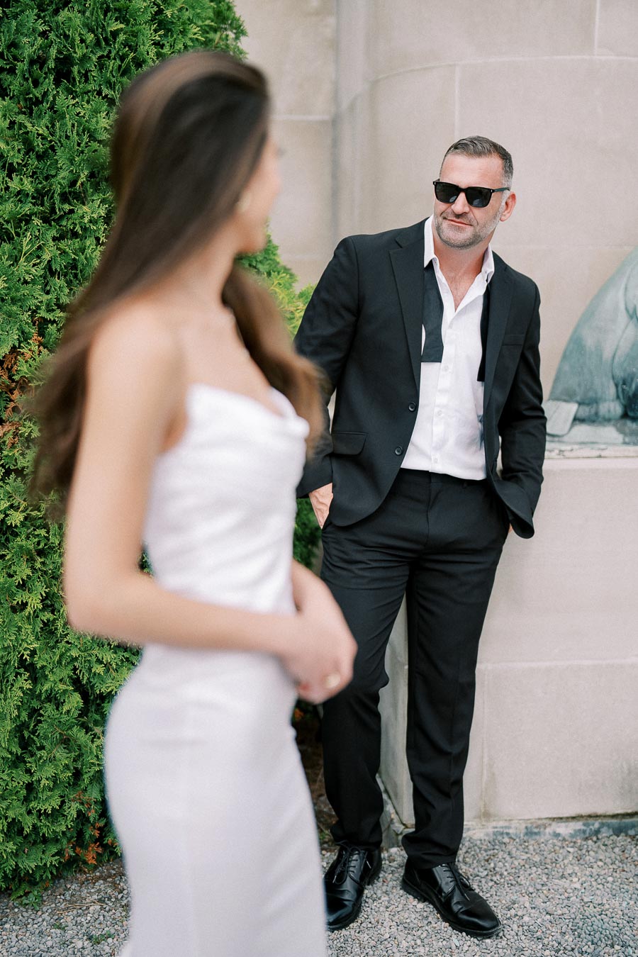 Man in a stylish black suit with sunglasses leaning against a stone wall, looking towards a woman in a white dress in an outdoor setting with greenery.