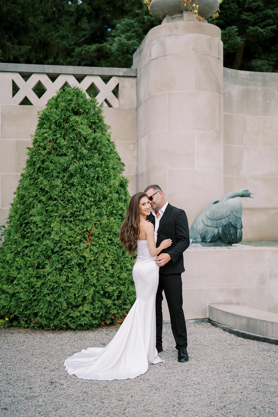 A couple in elegant attire embraces in a picturesque garden setting, with a lush green tree and decorative stonework in the background, ideal for a wedding or engagement setting.