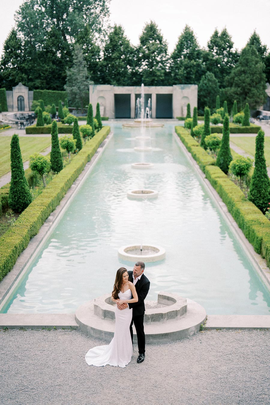 Elegant couple embracing near a beautiful, symmetrical garden fountain, with manicured greenery and classical architecture in the background.