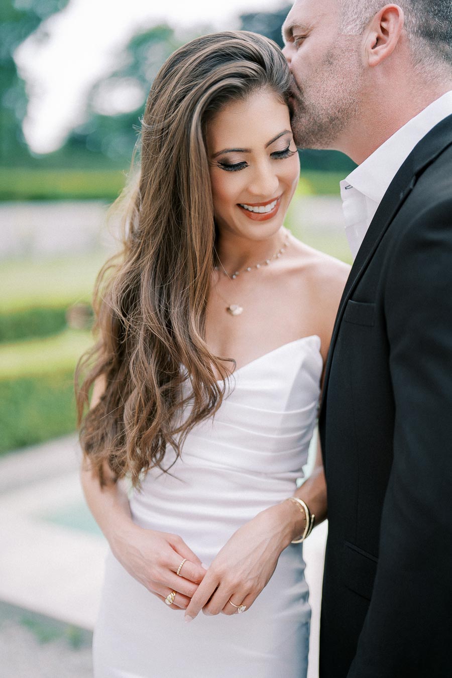 Couple sharing a romantic moment outdoors; woman in a white strapless dress smiling as man in a black suit gently kisses her head, surrounded by lush greenery.