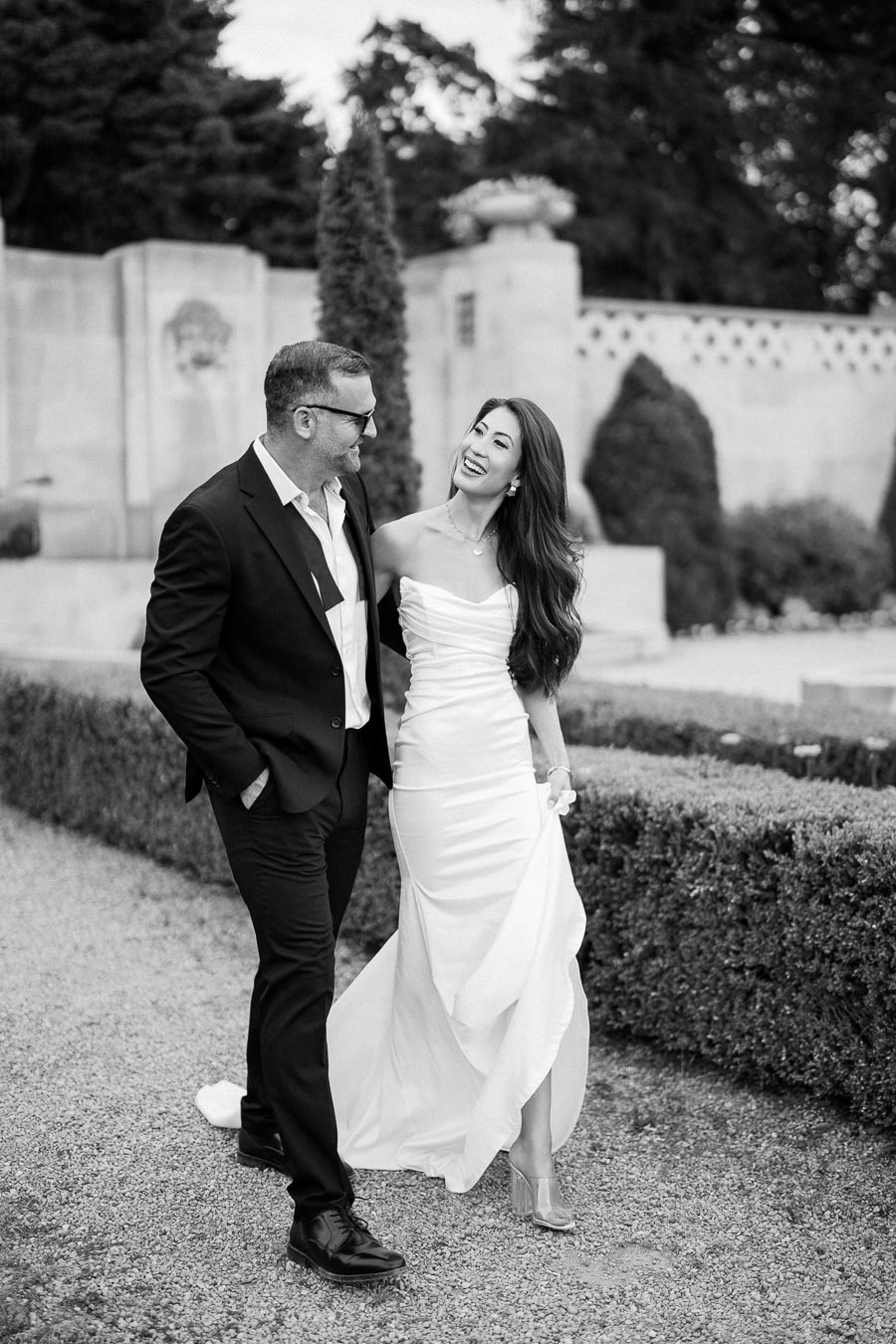 Black and white photo of a happy couple walking in a garden. The woman is wearing a strapless white dress, and the man is in a dark suit. They are smiling and looking at each other, surrounded by trimmed hedges and a stone wall backdrop.