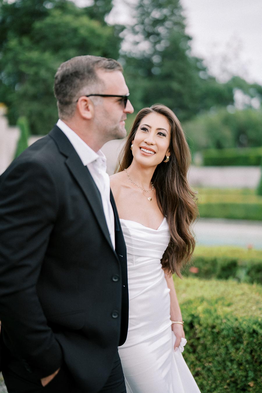 A woman in a white dress smiles at a man in a black suit with sunglasses, walking outdoors in a garden setting.