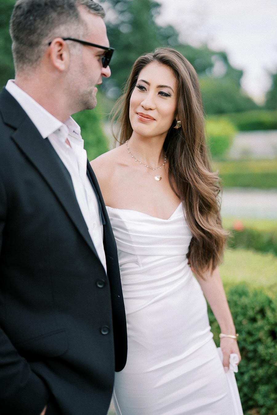 A couple in formal attire enjoying a moment together outdoors, with a woman in a white dress smiling at a man in sunglasses and a black suit jacket, surrounded by lush greenery.
