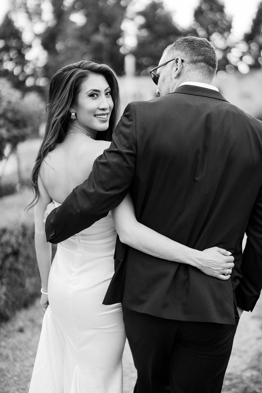 Black and white image of a smiling bride in a strapless gown walking arm in arm with a groom in a suit, both looking back over their shoulders in an outdoor setting.