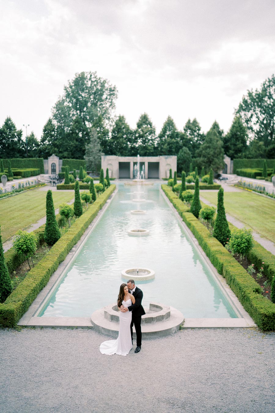A couple embraces by a picturesque reflecting pool in a formal garden setting, surrounded by manicured hedges and towering trees, with a stately stone facade in the background.