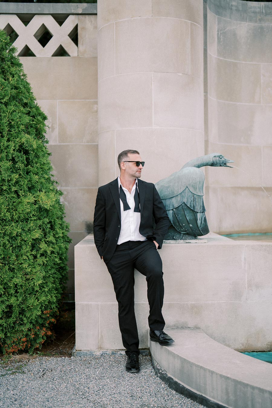 Man in a suit with an untied bowtie standing near a stone fountain, posing against a vintage architectural backdrop with greenery.