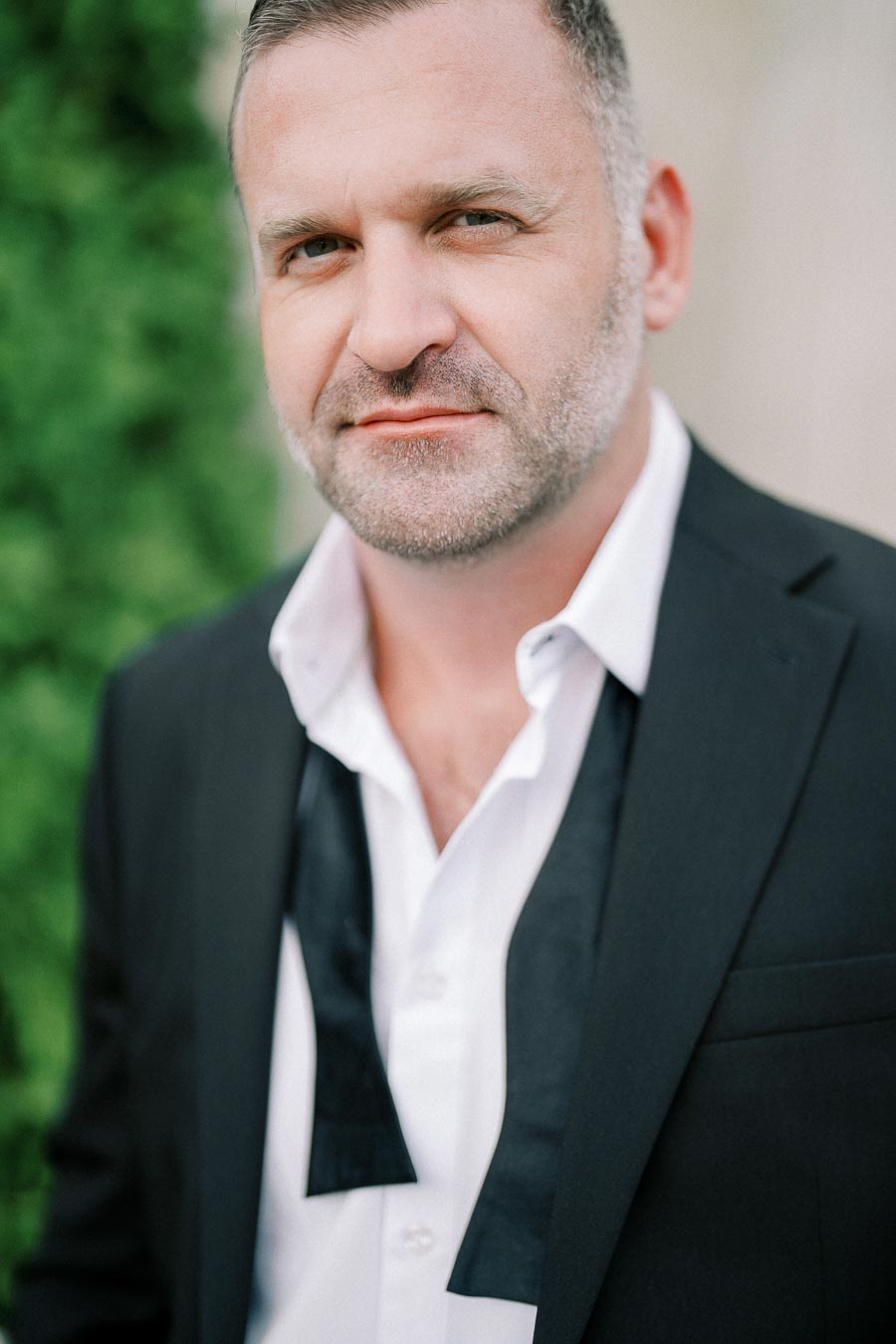 Portrait of a man in a formal black suit with an open collar and untied bow tie, standing outdoors with greenery in the background.