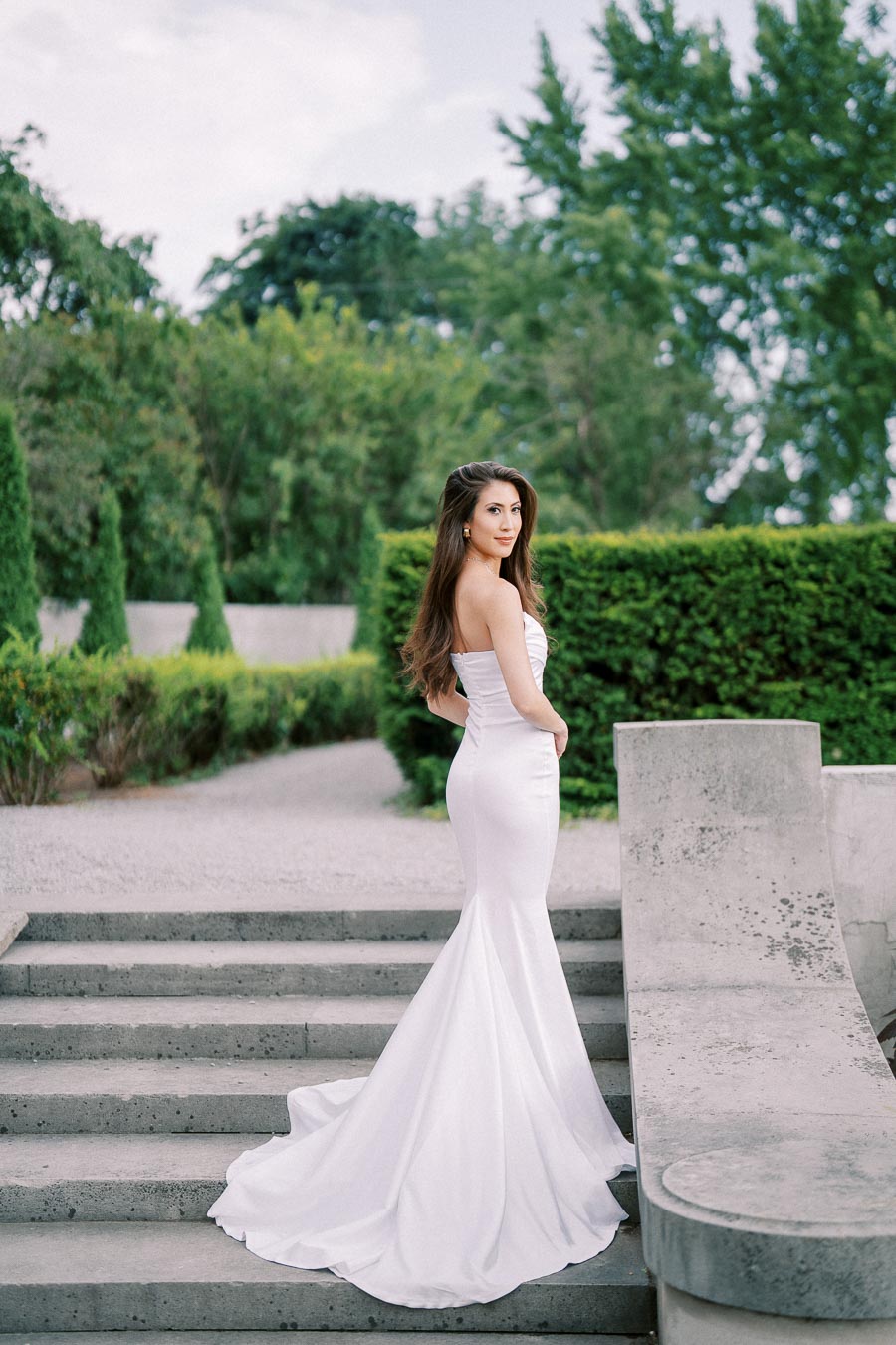 Elegant woman in a white wedding dress posing on outdoor stone steps surrounded by lush greenery.