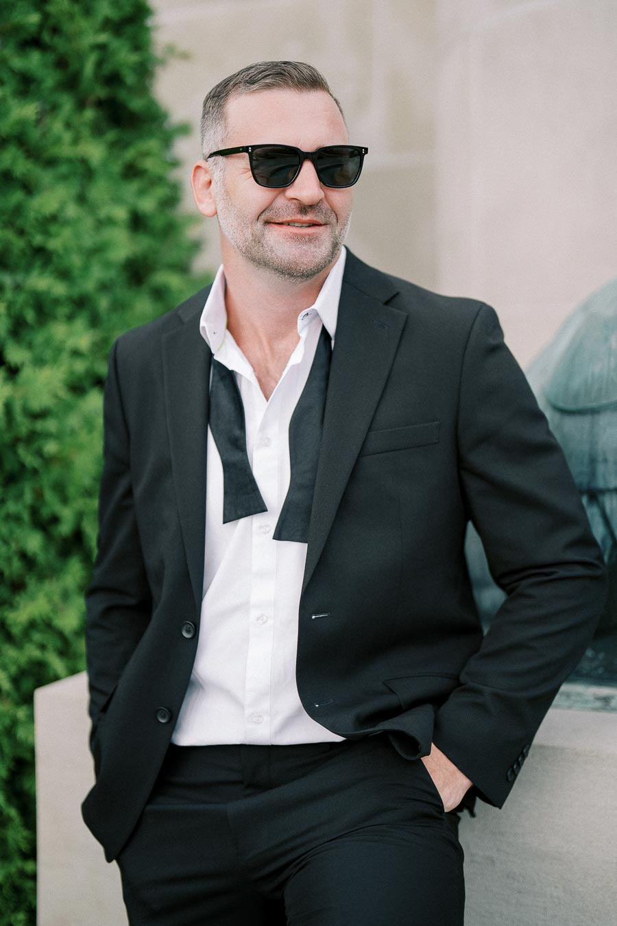 A smiling man in a stylish black suit with an open collar white shirt and untied bowtie, wearing sunglasses and leaning against a wall with greenery in the background.